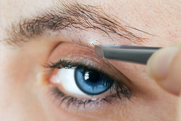 Close-up of a person shaping their eyebrow with tweezers, focusing on detailed eyebrow grooming.