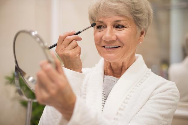 Senior woman applying makeup while looking at a handheld mirror.